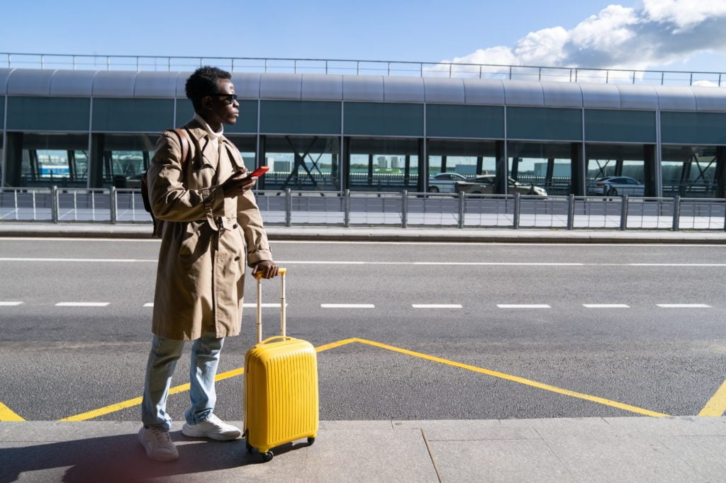 Black traveler man with suitcase stands in airport terminal holding phone calling looking for a taxi
